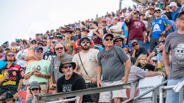 A large crowd of fans fills the Talladega Superspeedway grandstands, many wearing colorful shirts, hats, and sunglasses. They are smiling, waving, and cheering toward the camera.