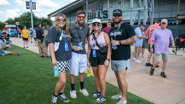A group of four people stands smiling on a grassy area at a racing venue, with the “Big Bill’s” building visible in the background. They are wearing casual summer clothes, sunglasses, and event lanyards, and are holding drinks. Other attendees walk and mingle in the background