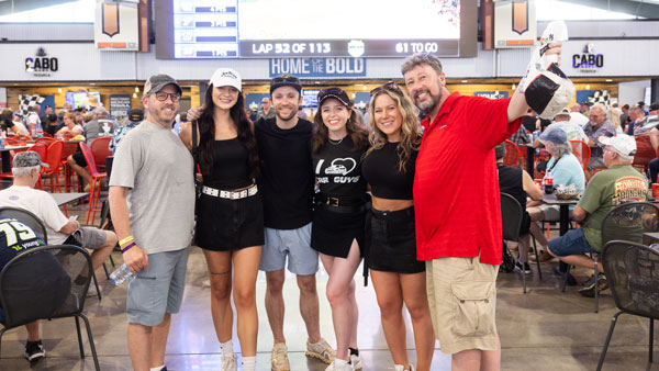 Six people pose together inside a busy indoor seating area at a racing event. They are smiling, some with arms around each other, wearing casual clothes, hats, and event wristbands. A large screen above them displays “Lap 52 of 113” and “Home of the Bold,” with many other attendees seated at tables in the background.