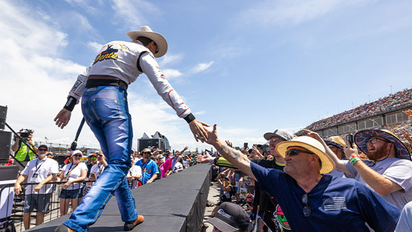 A driver wearing a cowboy hat, white shirt, and blue racing pants walks along a raised platform, reaching down to shake hands with a fan in a straw hat. A large crowd fills the grandstands and surrounds the stage under a bright blue sky.