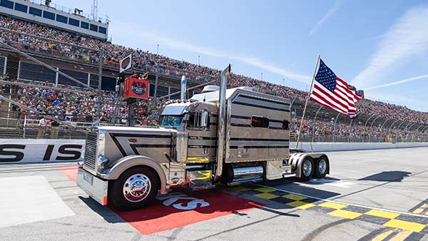 A polished silver semi-truck carrying a large American flag is parked on the start/finish line at Talladega Superspeedway. Packed grandstands fill the background as fans watch from the frontstretch under a clear blue sky.
