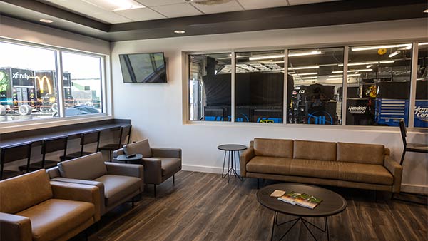 Modern climate-controlled garage suite with comfortable lounge seating, large viewing windows overlooking the NASCAR garage, high-top counter seating by the windows, wall-mounted televisions, and views of race team haulers and garages outside.
