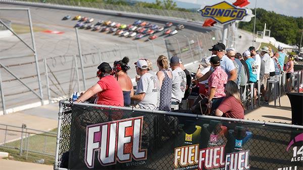 Fans watch a pack of NASCAR race cars speed around the track from the Turn 1 Deck hospitality area. Guests sit and stand along a railing overlooking the action, with FUEL by Franza branding on the deck and a Sunoco sign visible above the track.
