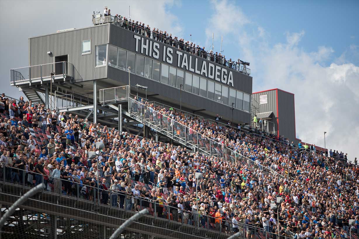 Wide view of Talladega Superspeedway with NASCAR race cars on track and packed grandstands surrounding the speedway.